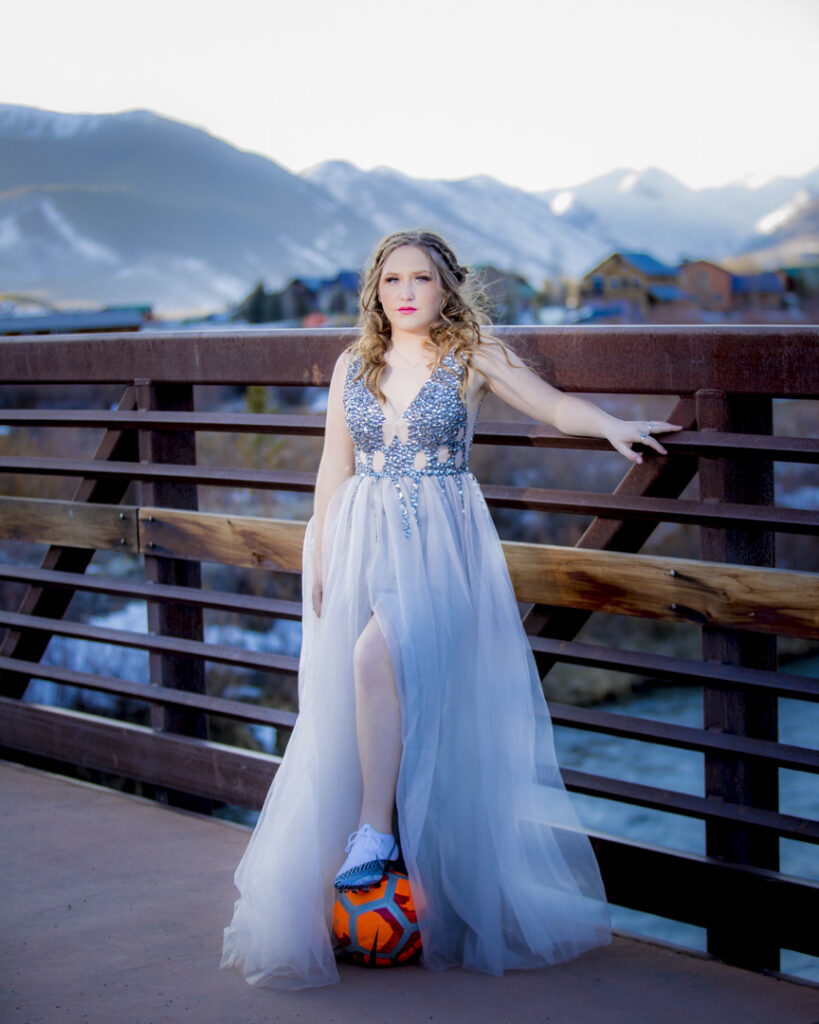 Portrait of a woman in a sparkling blue gown resting one hand on a bridge railing, foot on an orange soccer ball, with mountains in the background.