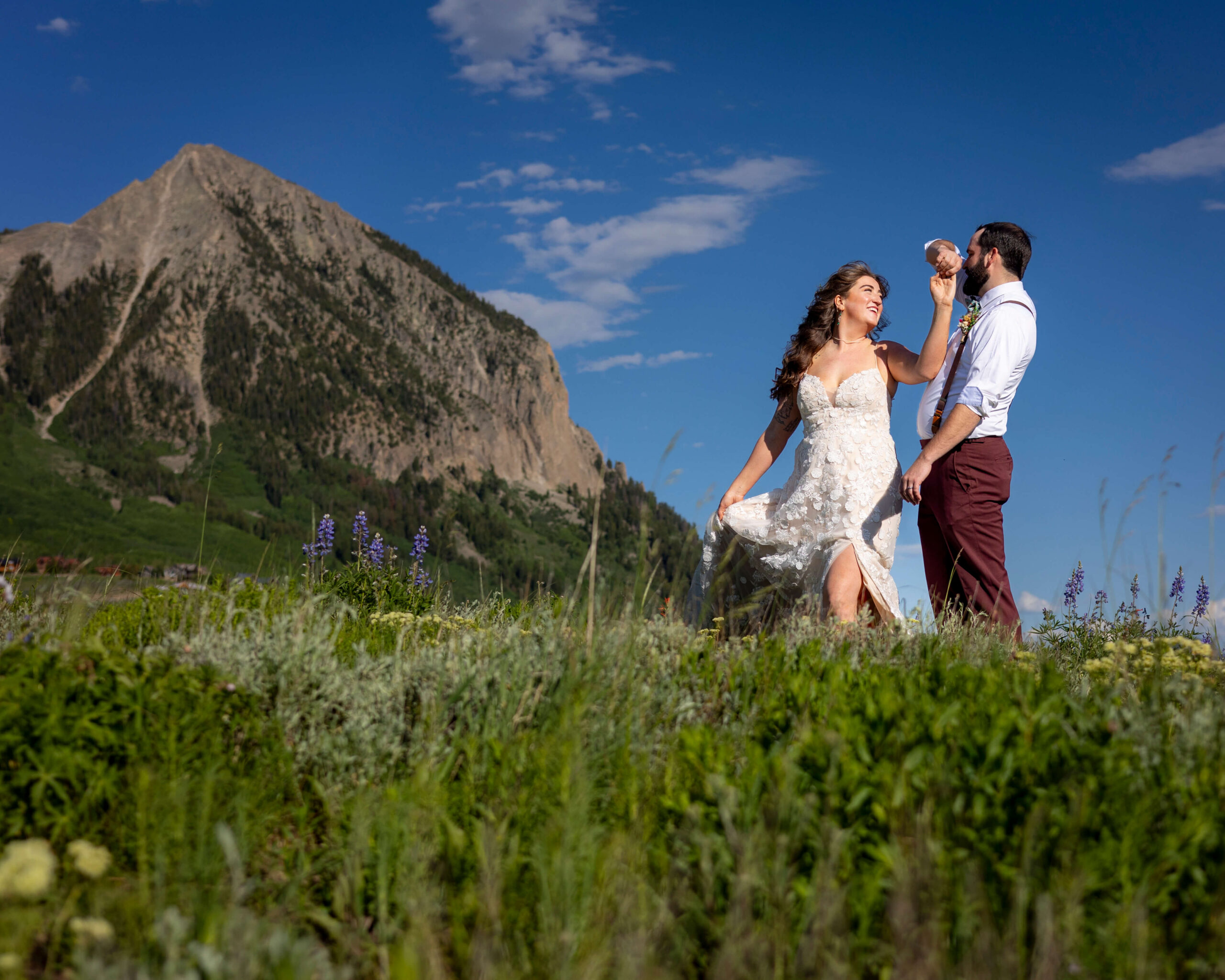 Bride in a lace wedding dress and groom in suspenders stand in a grassy meadow with wildflowers, mountains in the background under a clear blue sky.