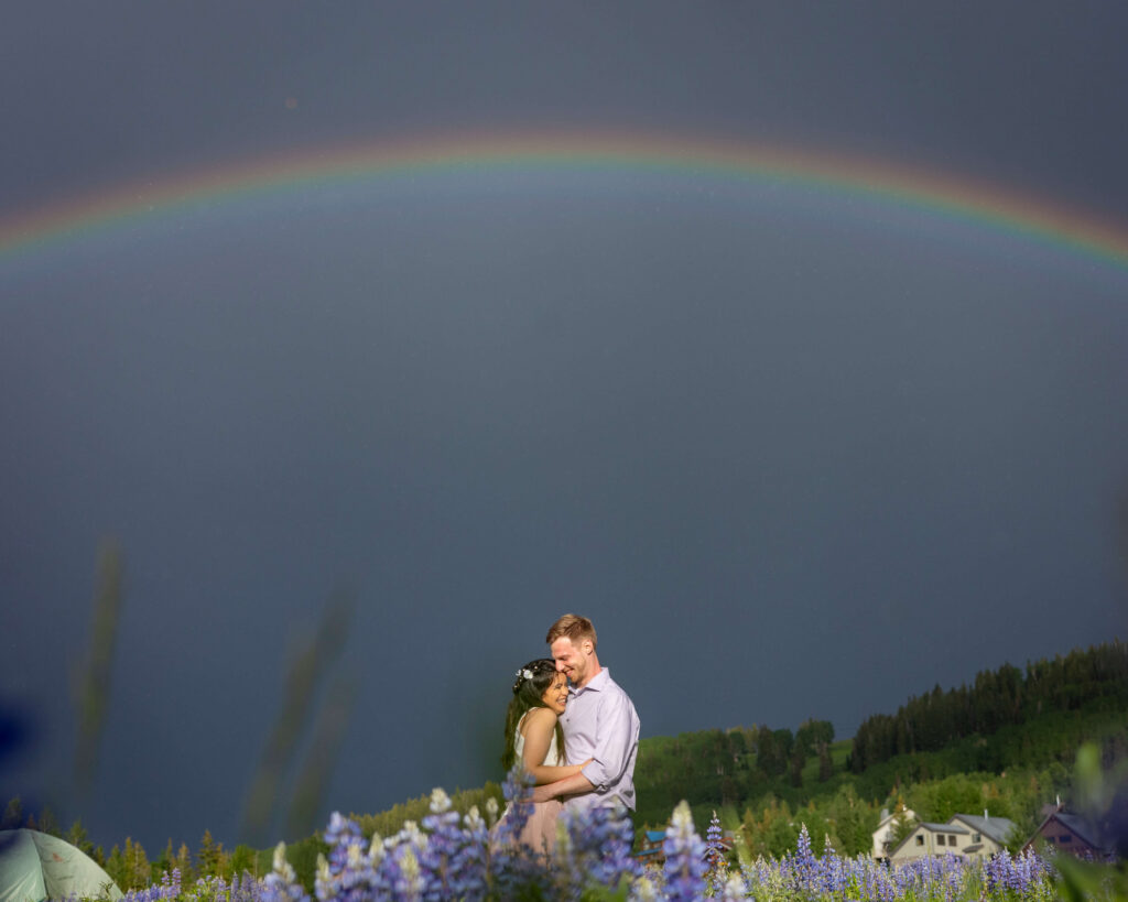Couple embraces in a meadow of purple flowers as a rainbow arcs across a clear sky.