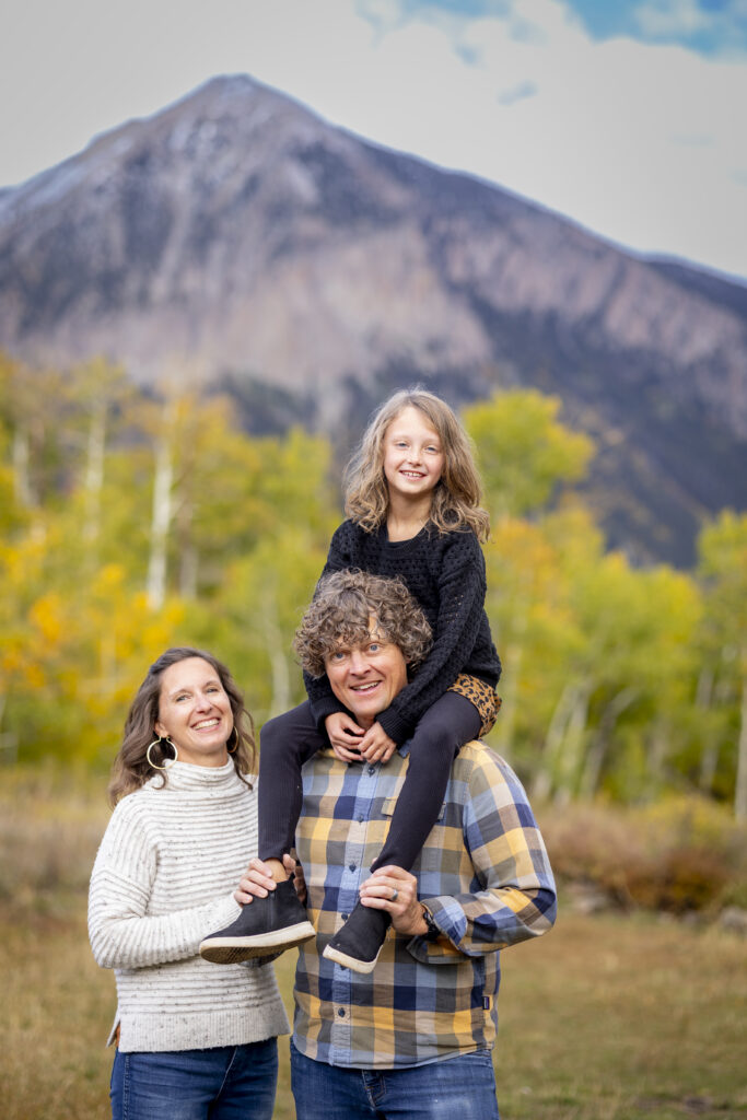 Smiling family outdoors in a field with mountains in the background; father carries daughter on his shoulders while mother stands beside them.