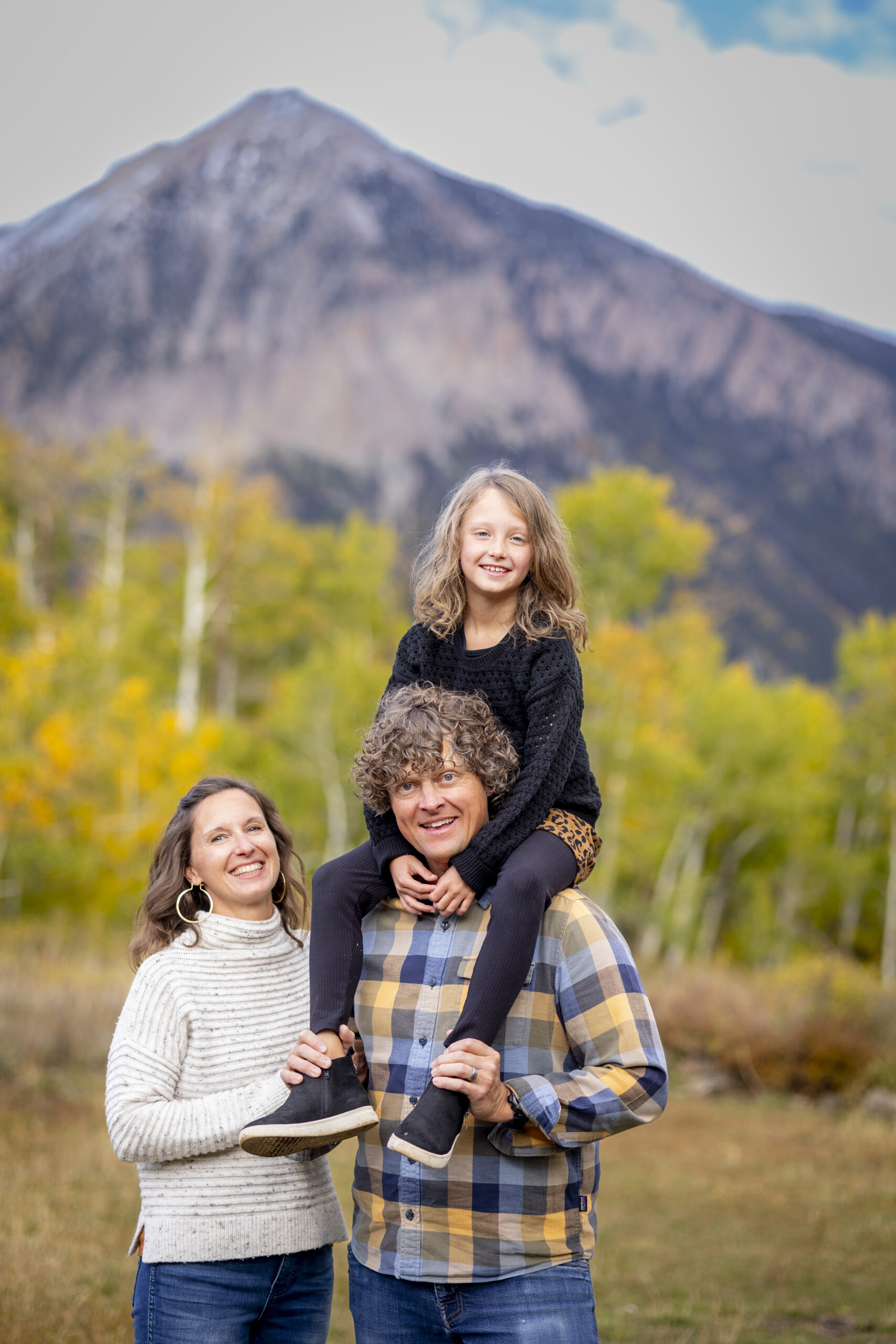 Smiling family outdoors in a field with mountains in the background; father carries daughter on his shoulders while mother stands beside them.