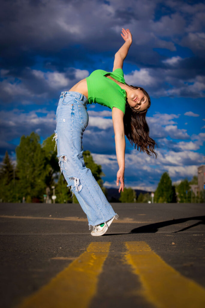 Woman in a bright green top and ripped jeans leans backward mid-air on a road with yellow lines, blue sky and clouds behind her for her senior pictures.