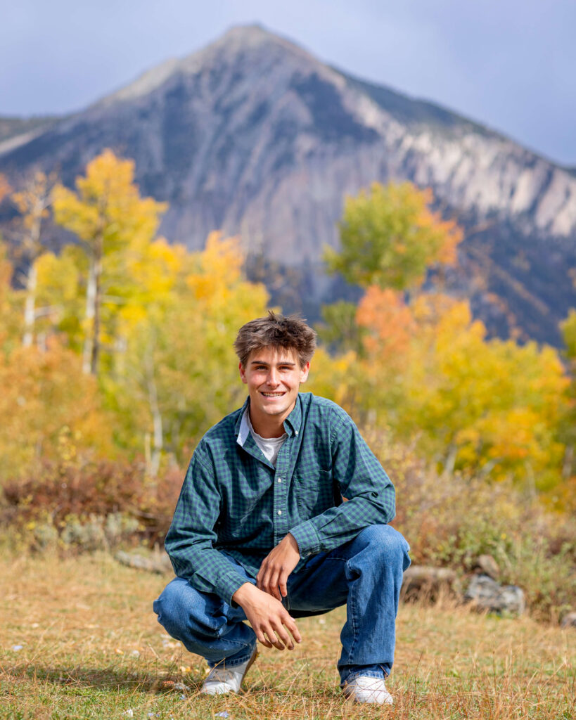 Young man crouching in a meadow with autumn trees and a distant mountain range behind him, smiling at the camera for his senior photography session.
