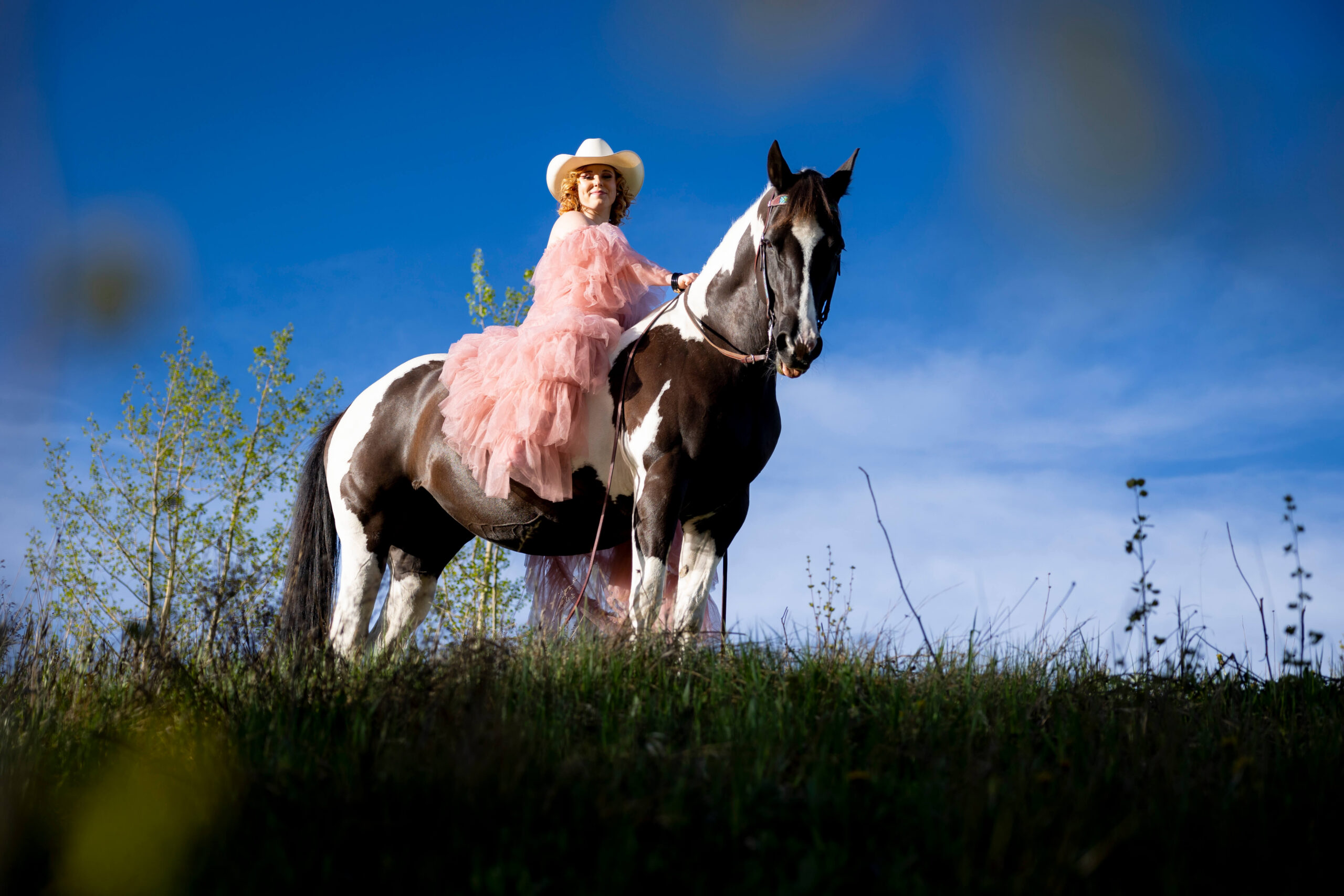 Woman in a pink ruffled dress and white cowboy hat rides a black-and-white horse across a grassy field under a clear blue sky.