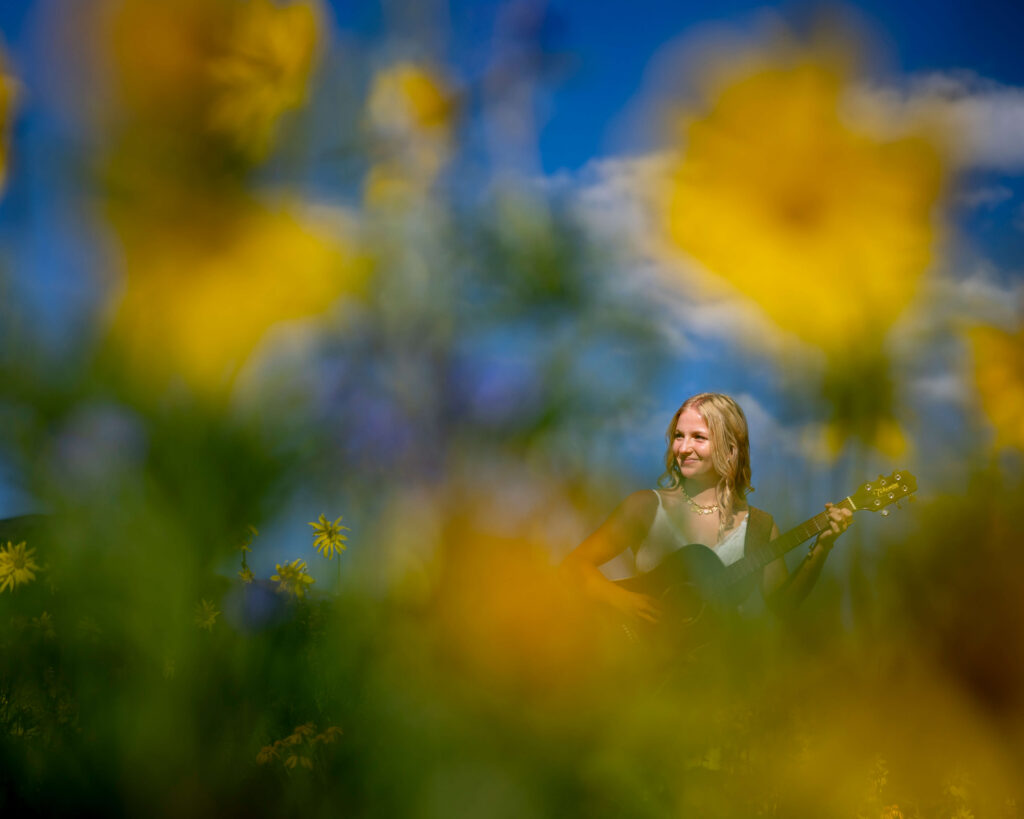 Person playing a guitar outdoors in a vibrant, blurred garden with yellow flowers and a blue sky background for her senior photos.