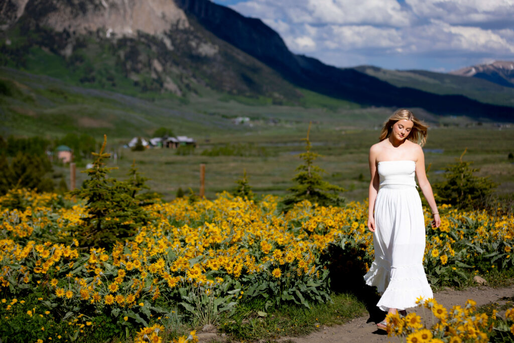 Woman in a white strapless dress standing among a sea of yellow flowers with mountains in the background for her senior photos in Crested Butte.