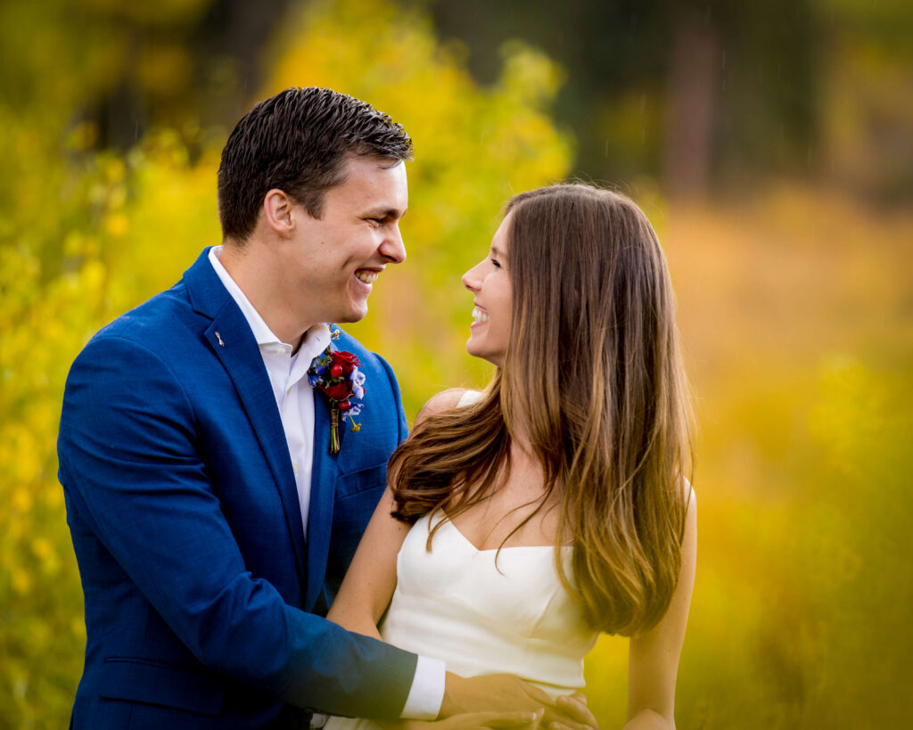 Couple embracing in a sunlit field, smiling at each other with yellow autumn foliage in the background.