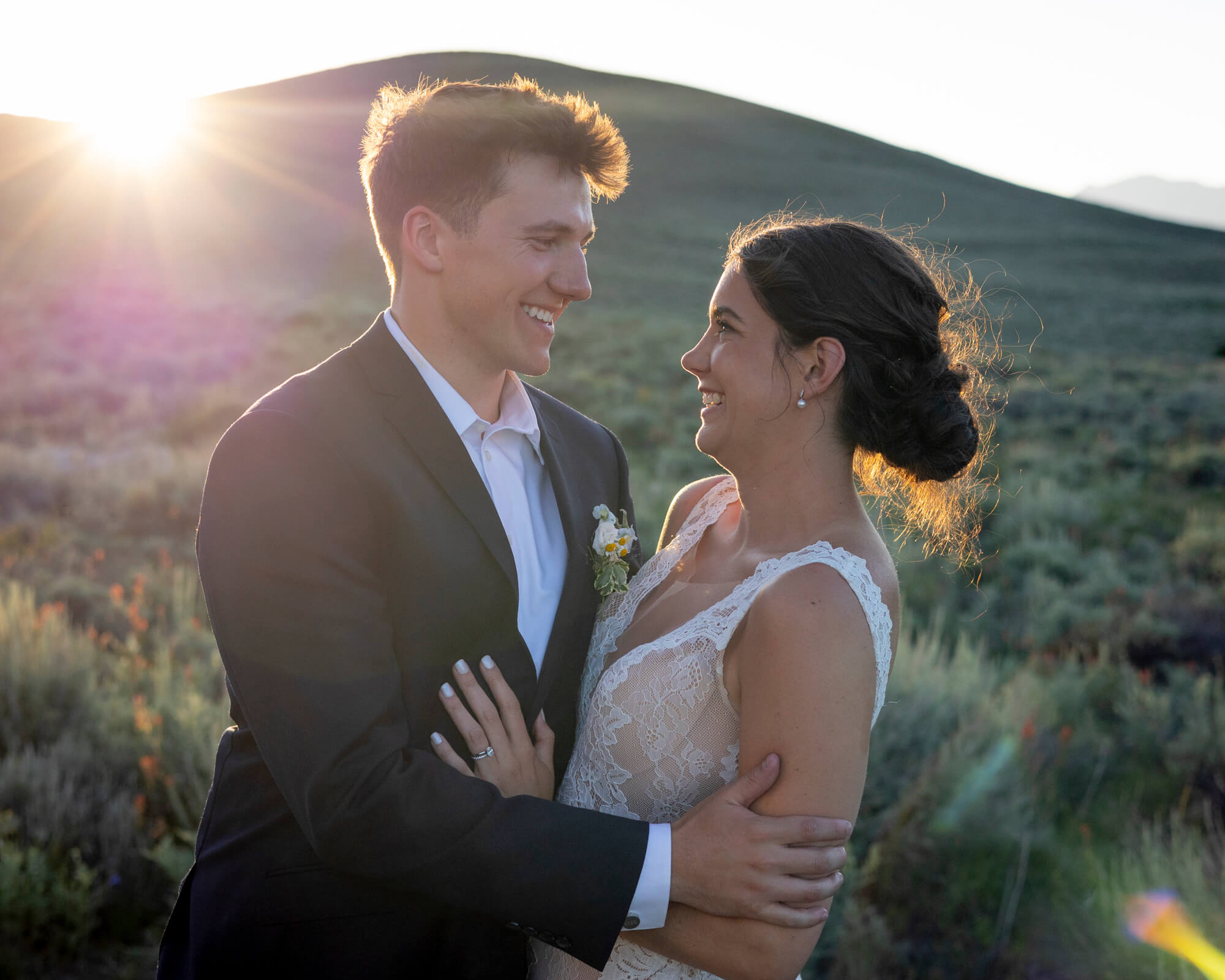Bride and groom embrace outdoors at sunset, smiling at each other with a sun flare behind them.