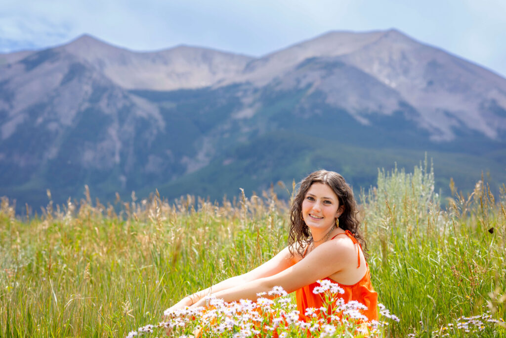 Smiling woman in an orange dress sitting in a meadow of wildflowers with mountains in the distance behind her for her senior photos in Crested Butte