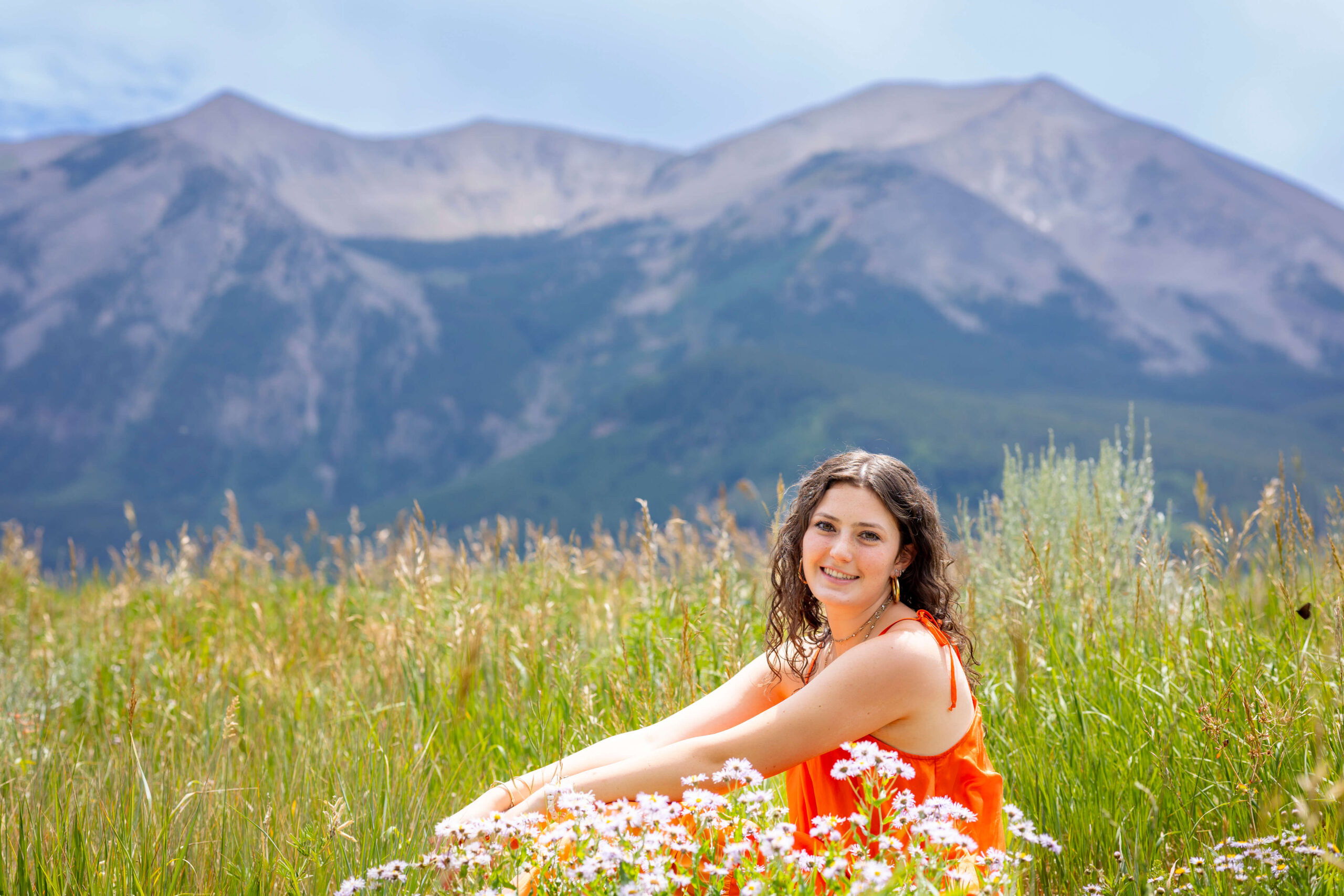Smiling woman in an orange dress sitting in a meadow of wildflowers with mountains in the distance behind her
