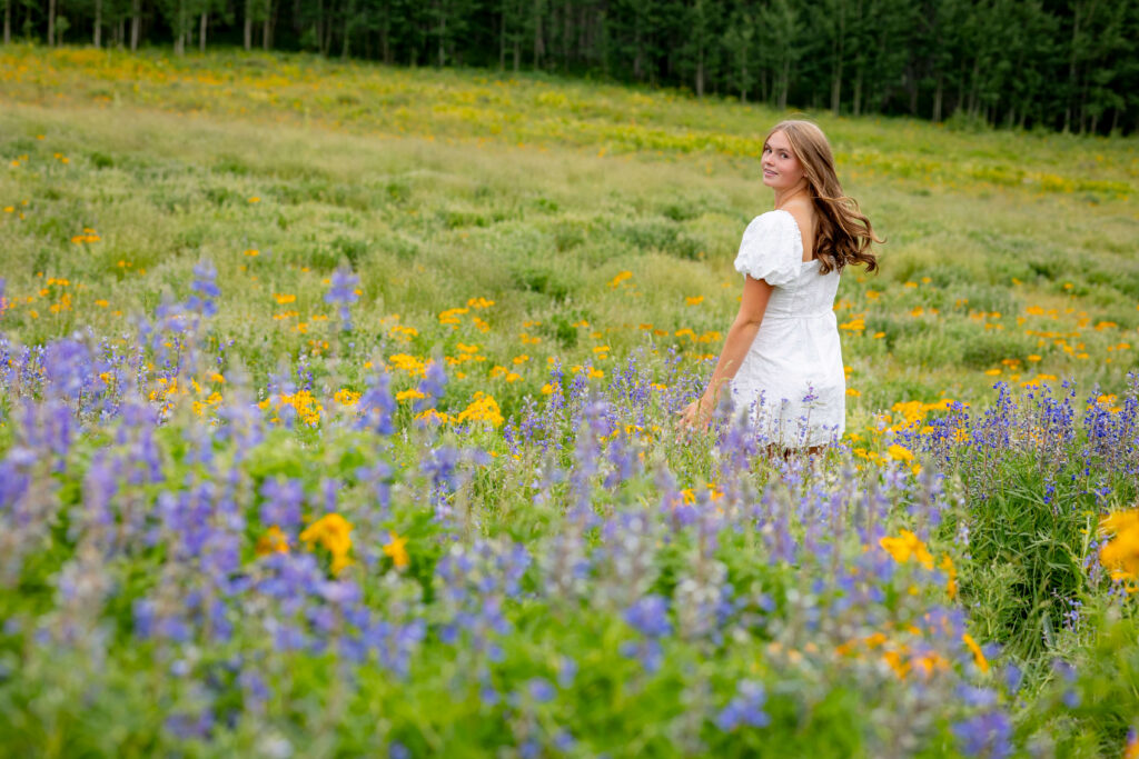 Woman in a white dress standing in a field of yellow and purple wildflowers with a forest in the background for her senior photos